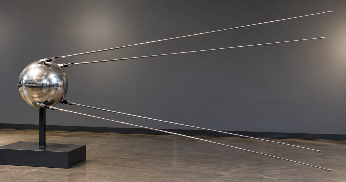 AI-generated photo-style museum display of a Sputnik 1 replica with a silver sphere and four long antennas